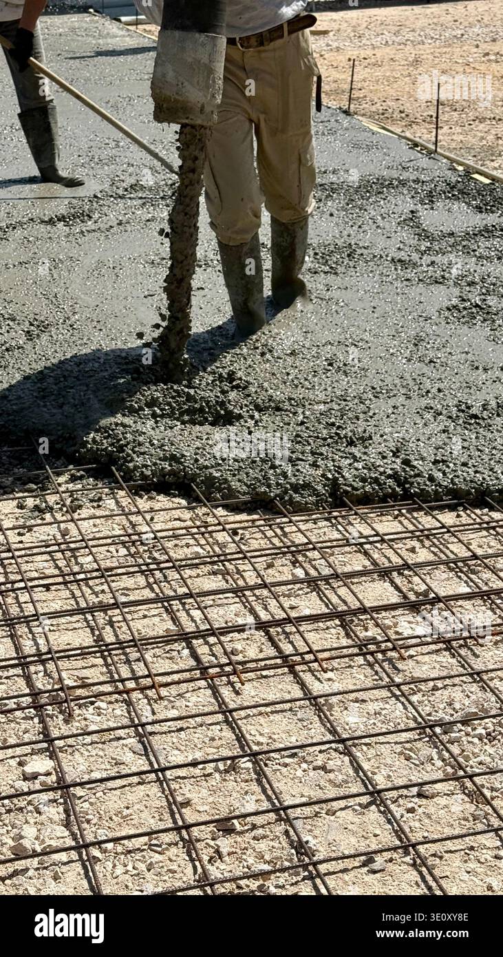 Nahaufnahme eines nassen Betons, der aus einem Pumpenschlauch auf ein Stahlbewehrungsgitter gegossen wird, mit den Stiefeln eines Bauarbeiters auf einer Baustelle. - Smartphone-aufgenommenes Stockfoto