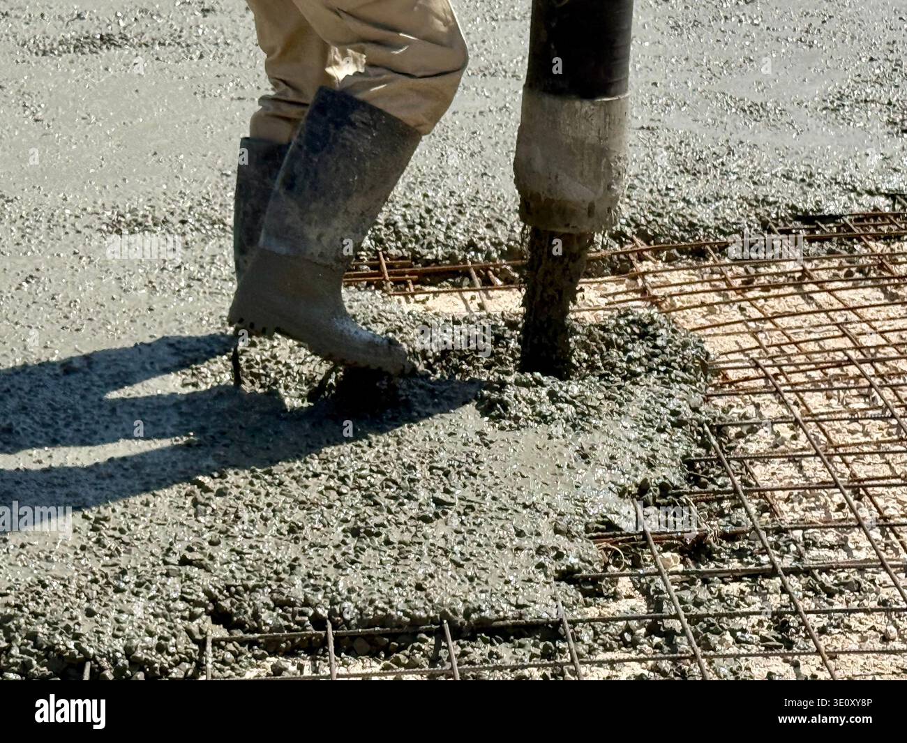 Nahaufnahme eines nassen Betons, der aus einem Pumpenschlauch auf ein Stahlbewehrungsgitter gegossen wird, mit den Stiefeln eines Bauarbeiters auf einer Baustelle. - Smartphone-aufgenommenes Stockfoto