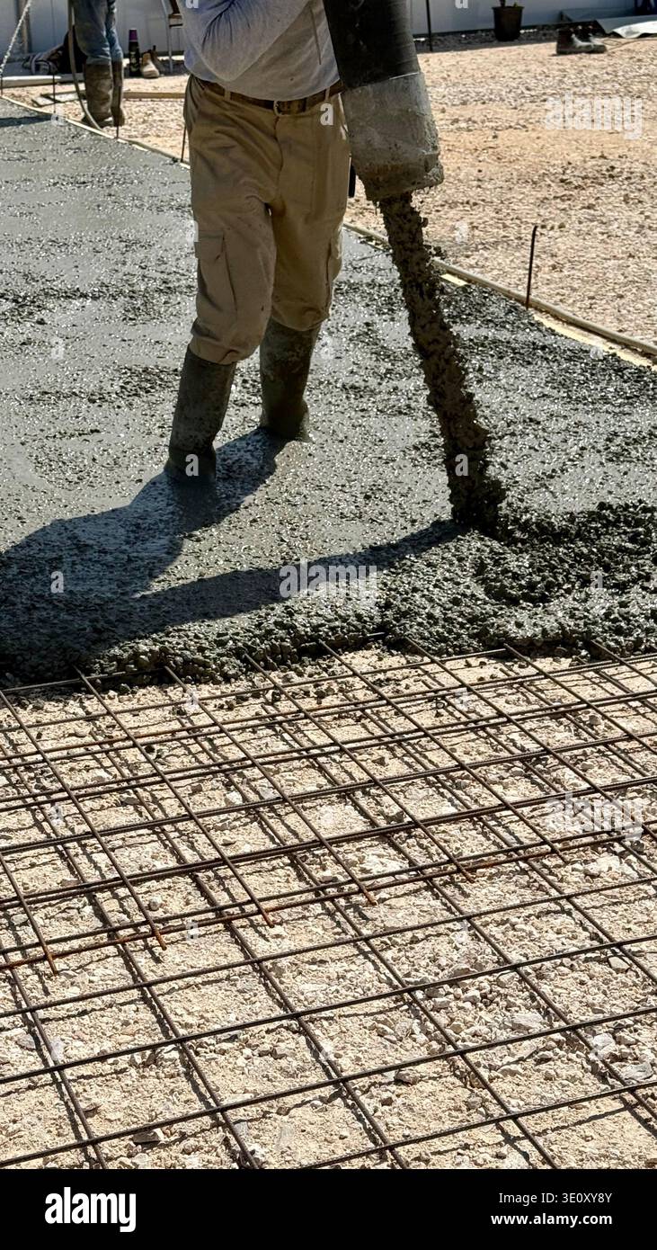 Nahaufnahme eines nassen Betons, der aus einem Pumpenschlauch auf ein Stahlbewehrungsgitter gegossen wird, mit den Stiefeln eines Bauarbeiters auf einer Baustelle. - Smartphone-aufgenommenes Stockfoto