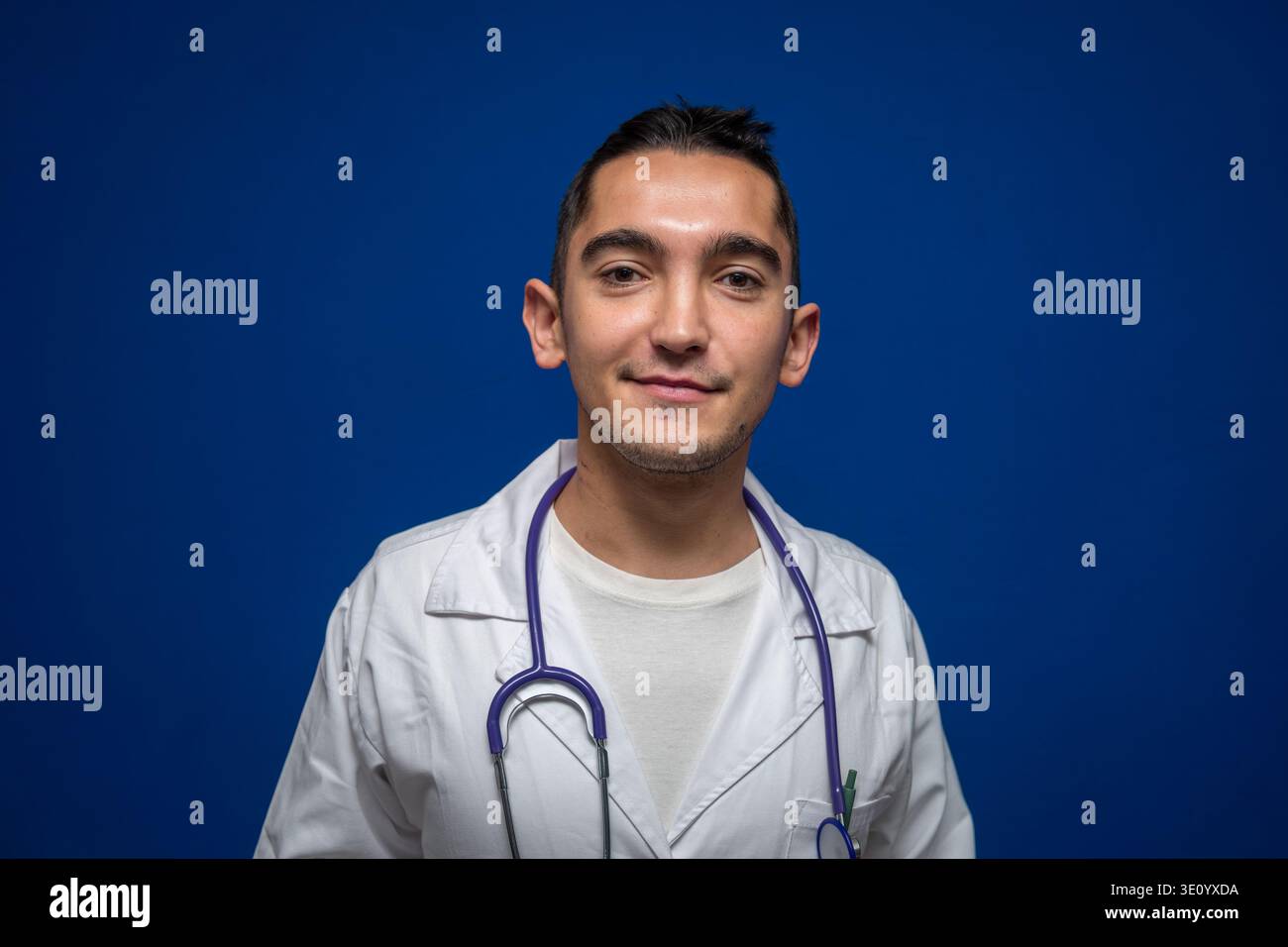 Junger Arzt mit weißem Mantel und lächelndem Stethoskop, stehend auf blauem Hintergrund, repräsentiert Gesundheit und Medizin Stockfoto