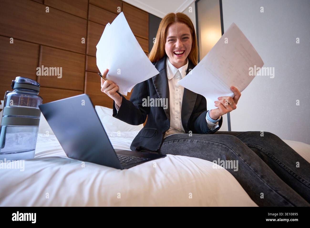 Junge Frau, die aufgeregt im Schlafzimmer mit Laptop und Papieren studiert Stockfoto