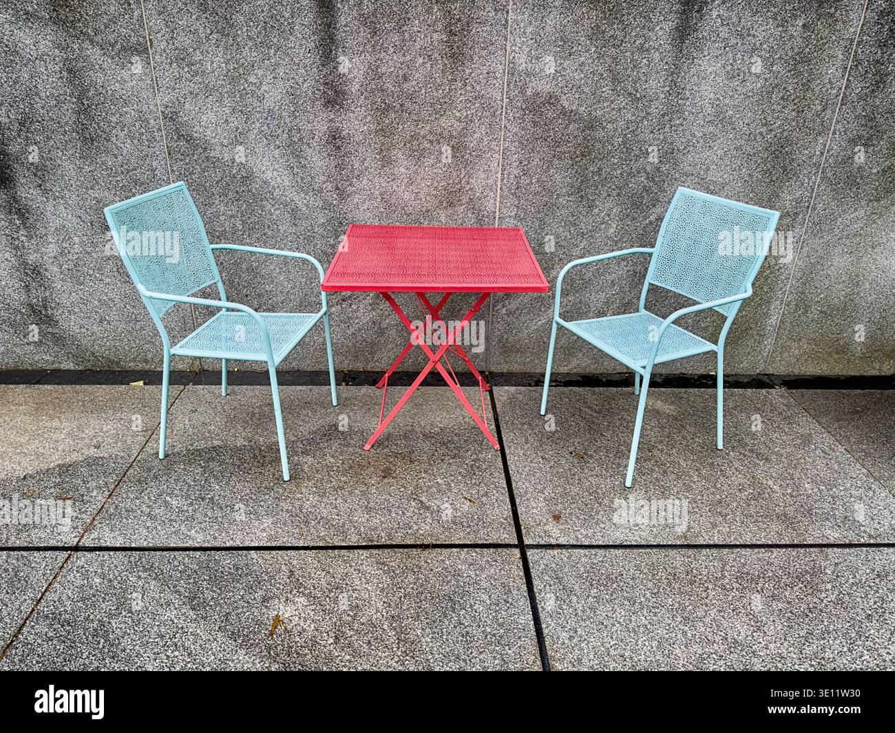 Gartenmöbel mit rotem Tisch und blauen Stühlen verleihen dem Innenhof aus Beton Farbe, die ansonsten fad wäre. Stockfoto
