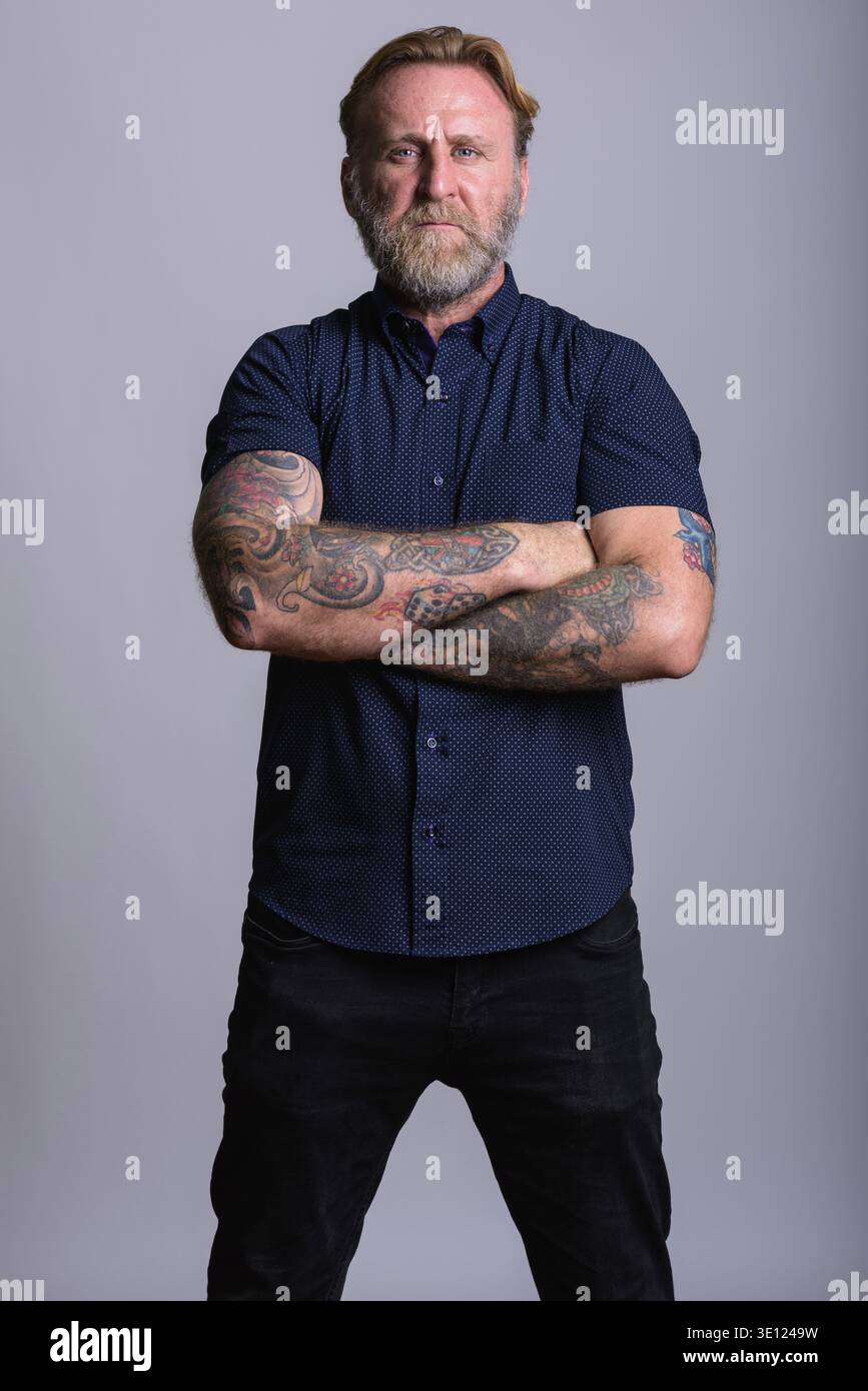 Studio shot reifer bärtiger Mann mit hand Tattoos stehend mit Waffen in grauer Hintergrund gekreuzt Stockfoto