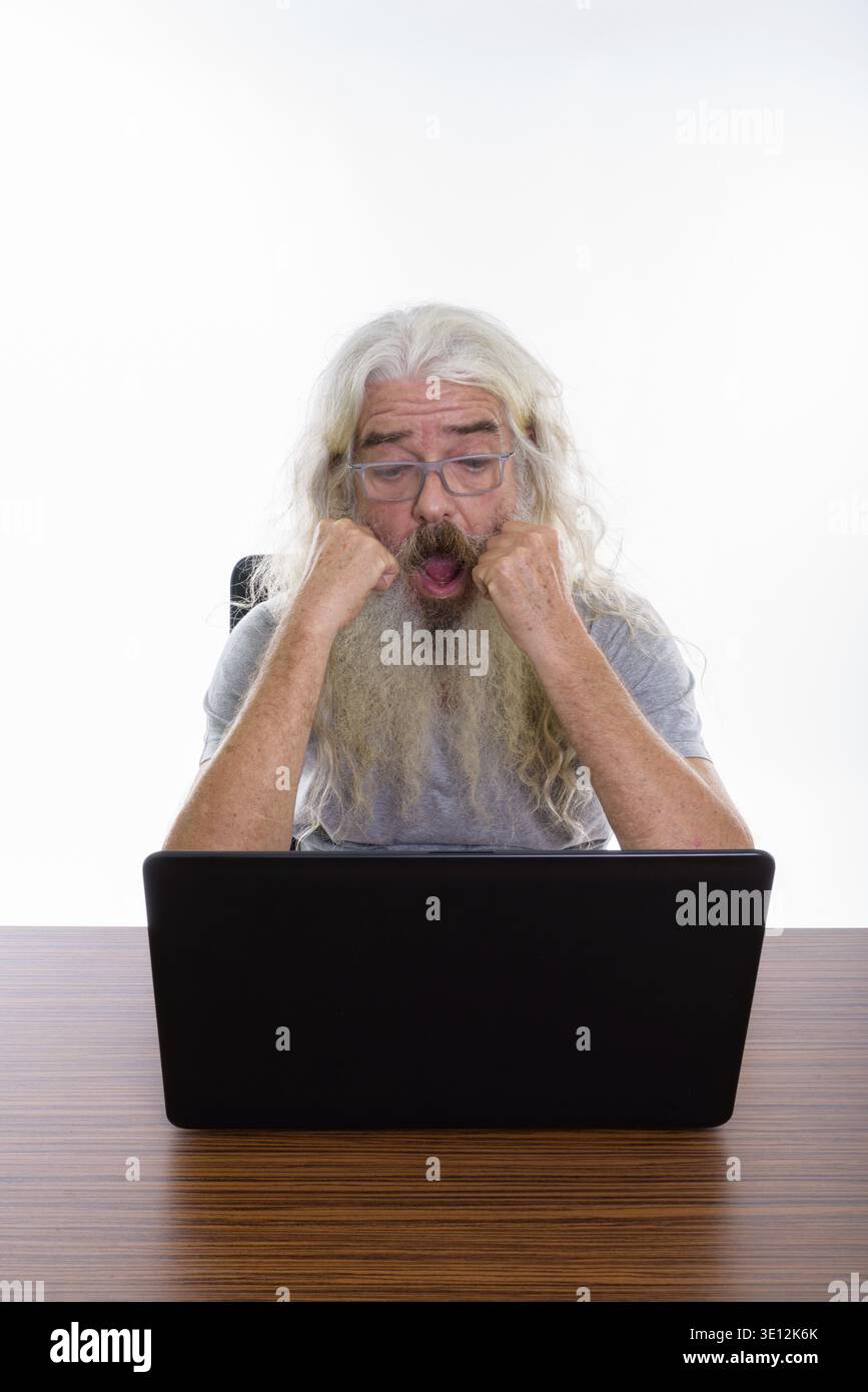 Studio shot der älteren bärtigen Mann Brille tragen, während Sie schockiert und mit Laptop auf hölzernen Tisch Stockfoto