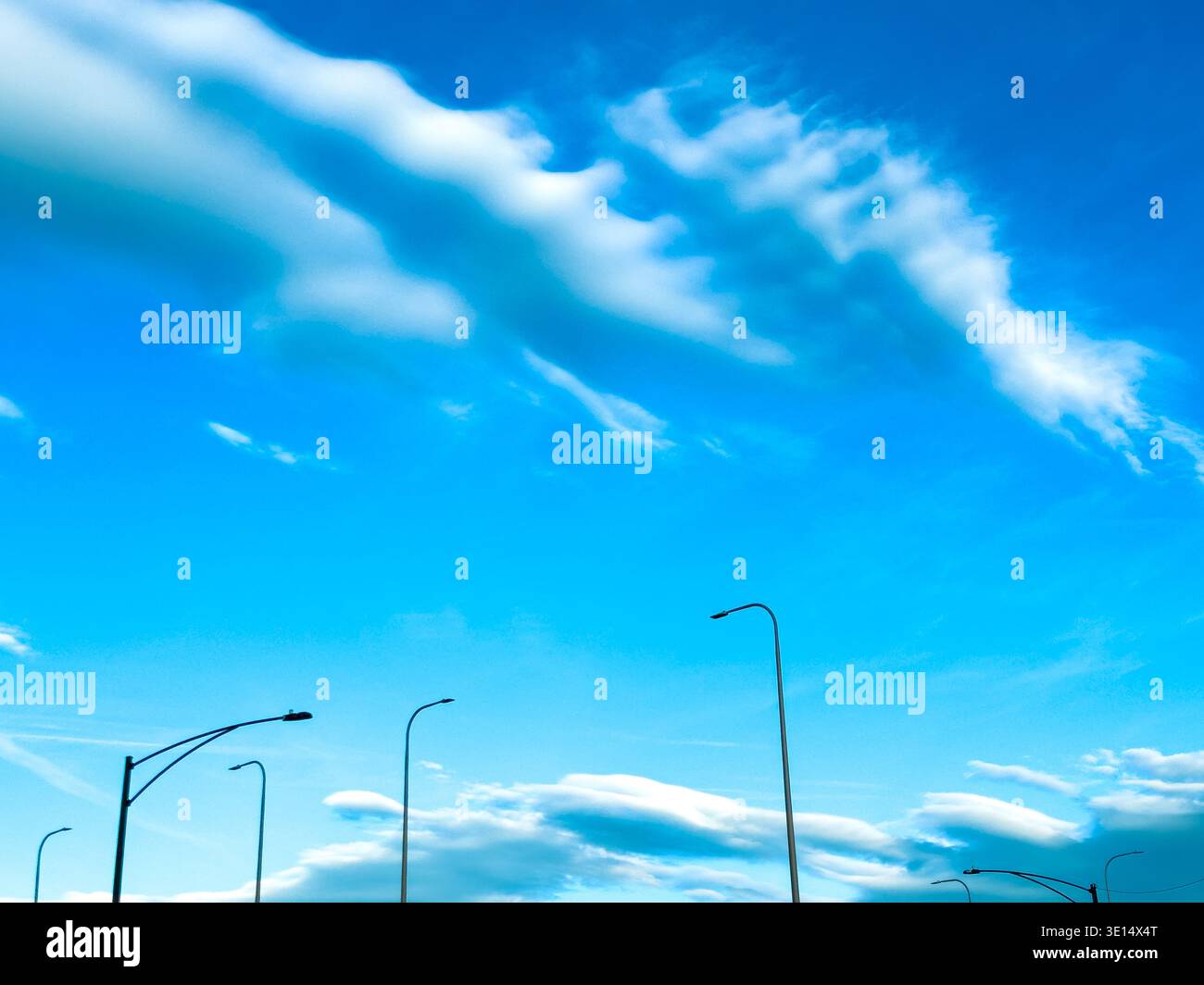 Weiße Wolken bewegen sich langsam in einem blauen Himmel über einer Straße, die von Straßenlaternen gesäumt ist. Es ist tagsüber und die Szene zeigt eine klare und offene Sicht mit wenig Geburtshilfe Stockfoto