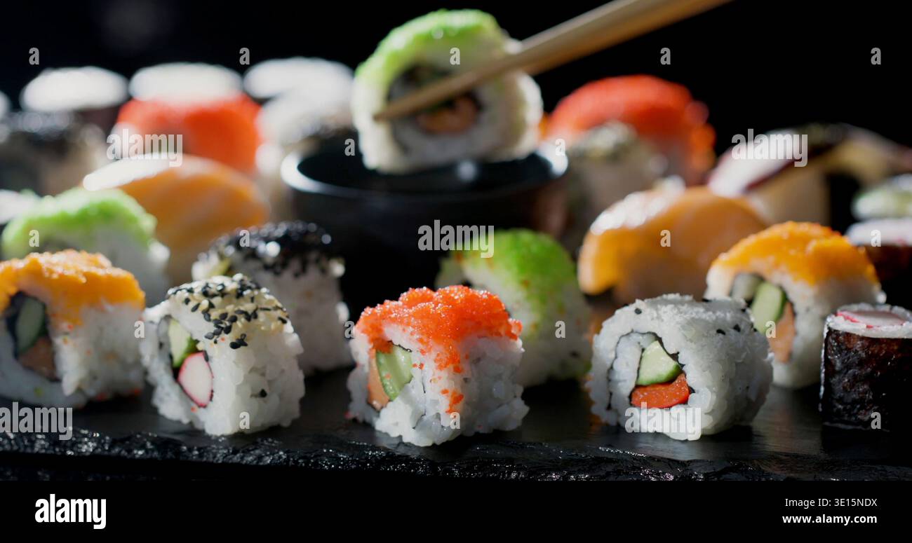 Verschiedene Sushi-Brötchen und Nigiri serviert auf einer schwarzen Platte mit Stäbchen, die ein Stück in einem Restaurant aufnehmen Stockfoto