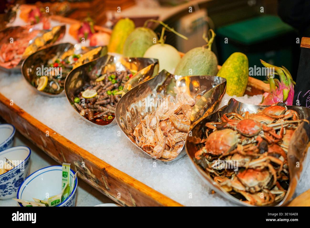 Frisches Meeresfrüchte-Buffet mit verschiedenen Krabben, Garnelen und Schnecken in großen, kühlenden Schüsseln, umgeben von Eis und exotischen Früchten wie Melonen und Stockfoto