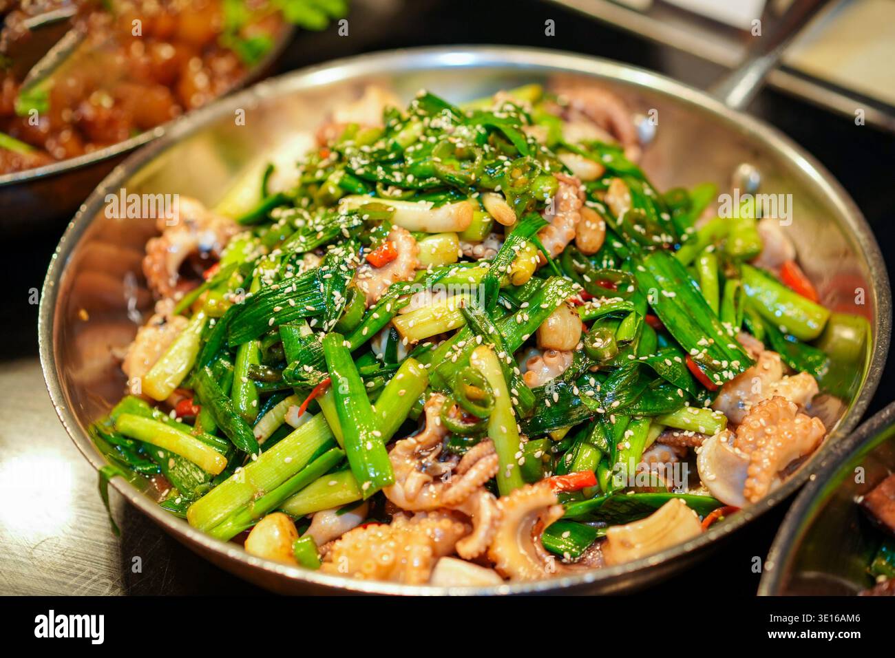 Tintenfisch mit leuchtendem grünen Knoblauch und roten Chili-Stücken, Braten in einer Metallpfanne, großzügig mit weißen Sesamsamen belegt, präsentiert eine Stockfoto
