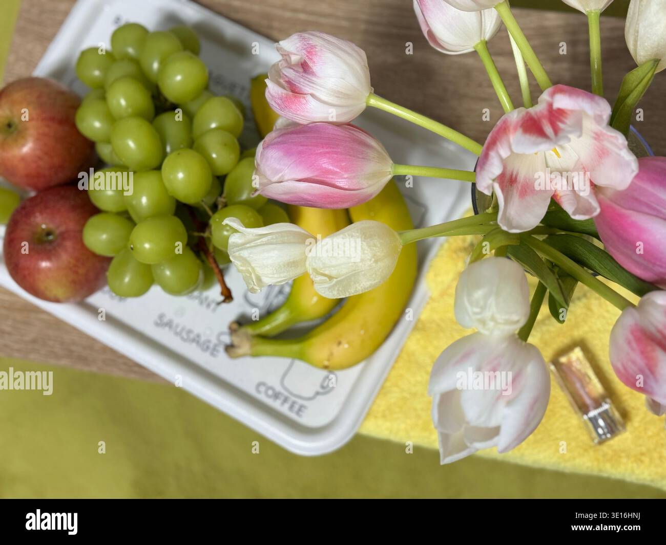 Tablett mit frischem Obst und grünen Trauben, Apfel, Bananen und Tulpenstrauß - Smartphone-aufgenommenes Stockfoto
