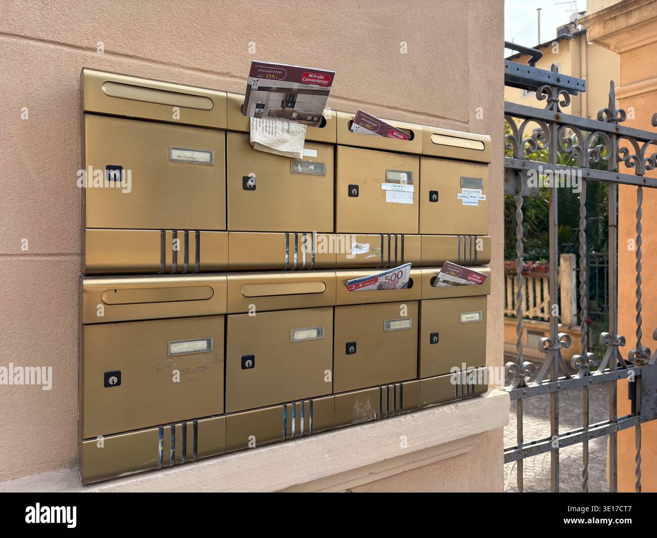 Ansammlung goldener Briefkästen mit Werbeblättern neben einem kunstvollen Metalltor, Pisa, Italien. Stockfoto