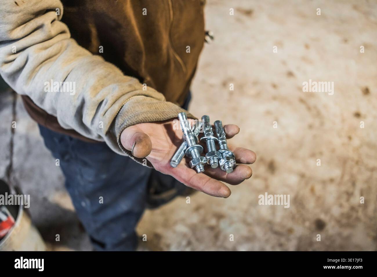 Ein Arbeiter in einer rauen Umgebung hält Metallbolzen in einer schmutzigen Hand und zeigt die Werkzeuge seines Fachs vor einem unscharfen industriellen Hintergrund Stockfoto