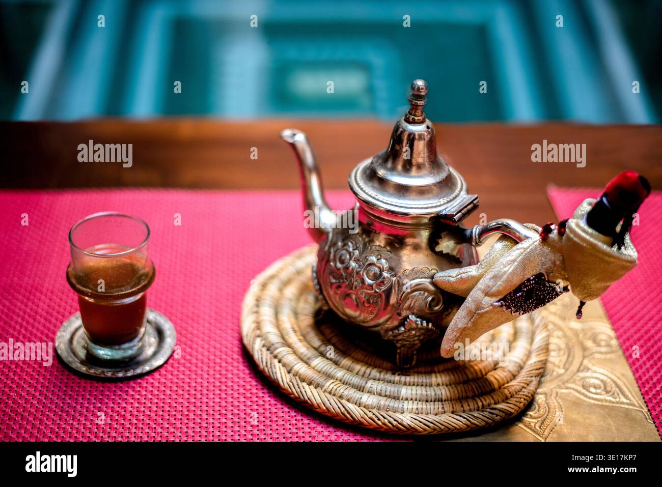 Dekorative traditionelle marokkanische Teekanne auf einer hellroten Matte, die an die Wärme von Marrakesch erinnert. Der aufwendig gestaltete Topf wird von begleitet Stockfoto