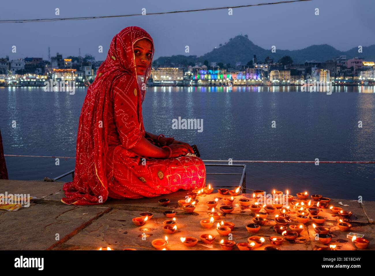 Eine junge Frau in einem roten Sari sitzt am Pushkar-See, Pushkar Sarovar, umgeben von beleuchteten Öllampen, wobei die beleuchtete Stadt in der Ferne sichtbar ist. Stockfoto