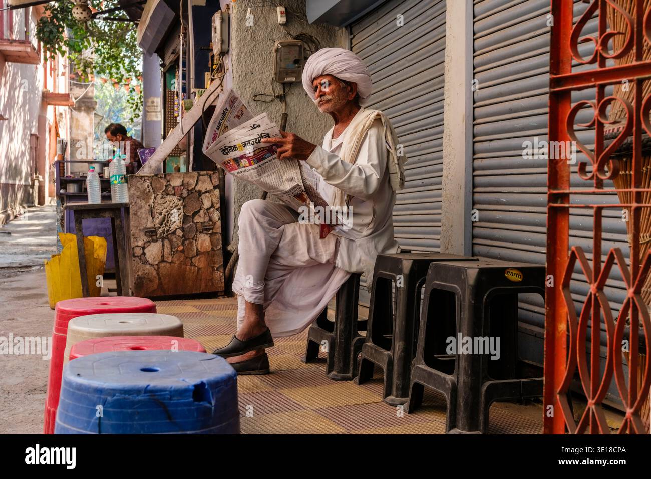 Ein Mann mit weißem Turban sitzt auf einem Plastikhocker in einer ruhigen Gasse von Puschkar, während er vor geschlossenen Ladenläden eine Zeitung liest. Stockfoto