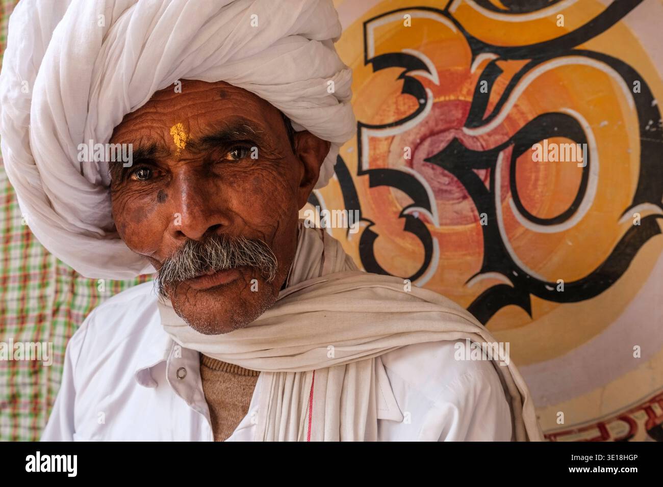 Ein Porträt zeigt einen älteren Mann in Puschkar, der einen großen weißen Turban und einen tilak auf der Stirn trägt und vor einem gemalten OM-Symbol posiert. Puschkar Ra Stockfoto