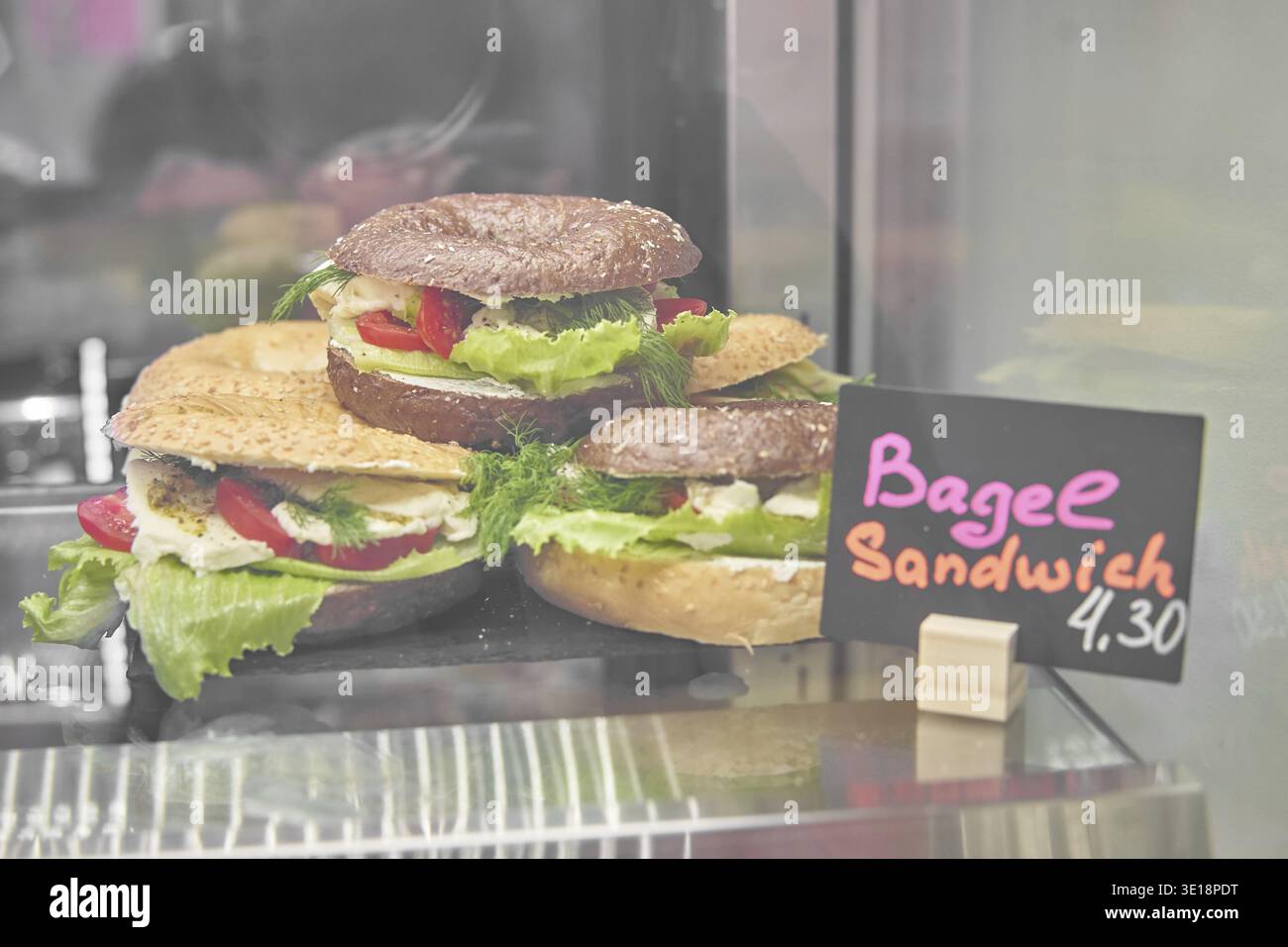 Drei Bagels mit Salat und Gemüse in einer Glasvitrine, Preisschild daneben, zum Verkauf in einer Bäckerei, einem Café, einem Pausenladen oder ca. Stockfoto