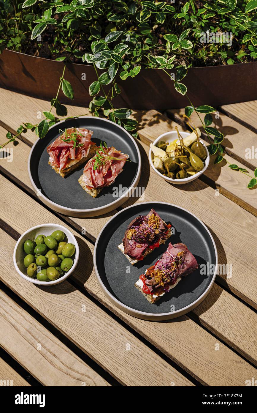 Auf zwei Tellern finden verschiedene Tapas mit Wurstwaren und Belägen sowie Schüsseln mit grünen und schwarzen Oliven in einer lebhaften Umgebung im Freien statt Stockfoto