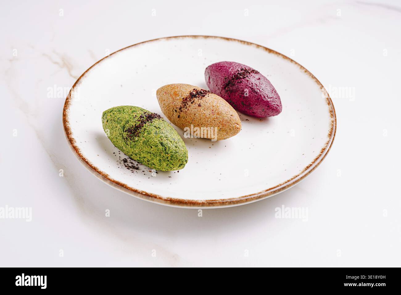 Ein Teller zeigt drei lebhafte Desserts in Form von kleinen Eiern. Die Aromen umfassen Matcha-Grüntee, Heidelbeere und Süßkartoffel, die jeweils leicht mit Staub bestäubt werden Stockfoto