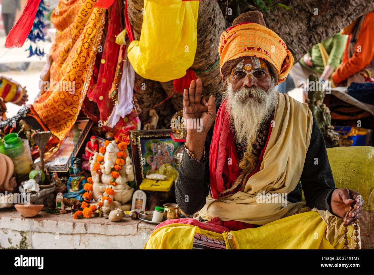 Bei Sunset Ghat am Pushkar Lake hebt ein Sadhu oder heiliger Mann mit Gesichtsmarkierungen seine Hand, während er neben einem Altar mit religiösen Figuren sitzt. Puschkar Stockfoto