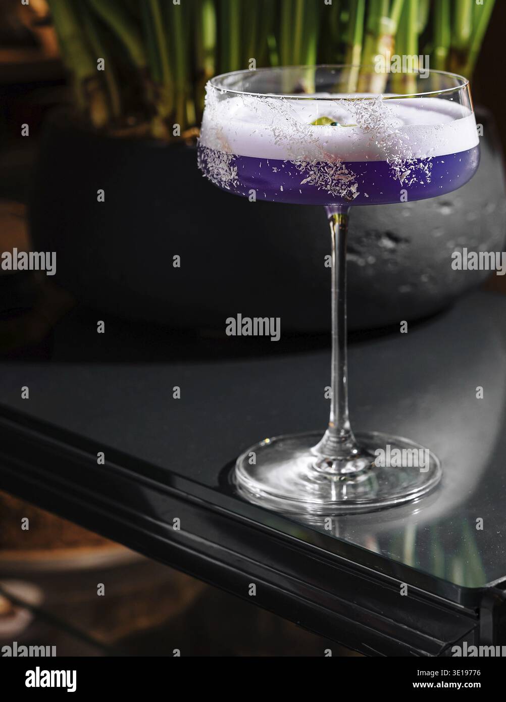 Lila Cocktail mit Eiweißschaum und Blumengarnitur auf einem Tisch mit einer Pflanze im Hintergrund Stockfoto