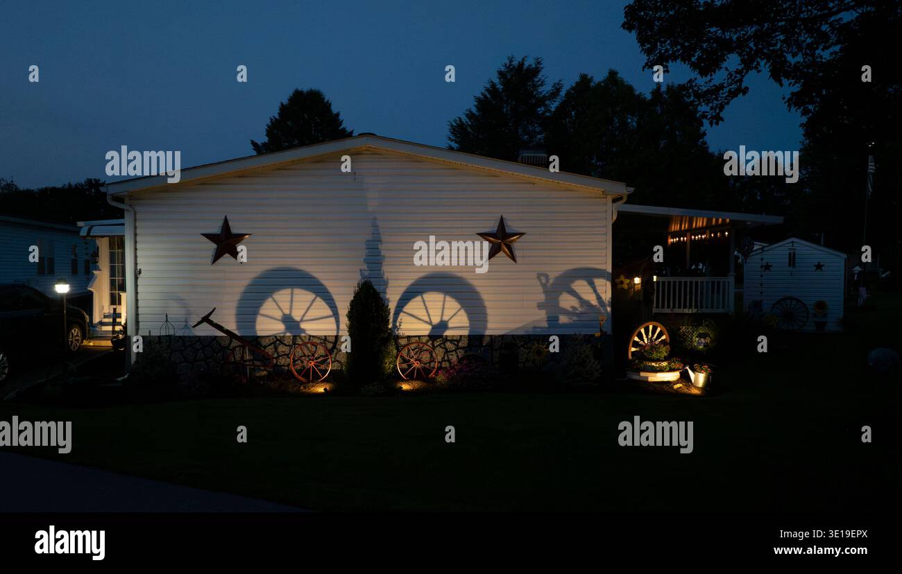 Lichter leuchten auf einem Mobilheim und erzeugen Schatten von Rädern und Sternen an der Wand. Die Szene zeigt einen ruhigen Abend. Das Haus ist von einem Garten mit einfachen Dekorationen umgeben. Stockfoto