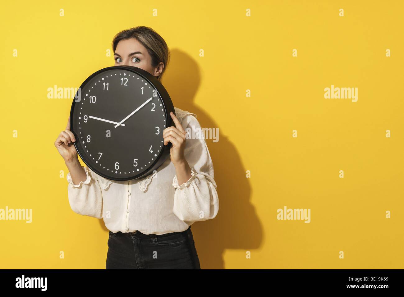 Eine junge Geschäftsfrau, die hinter einer großen Uhr auf gelbem Hintergrund rausblickt Stockfoto