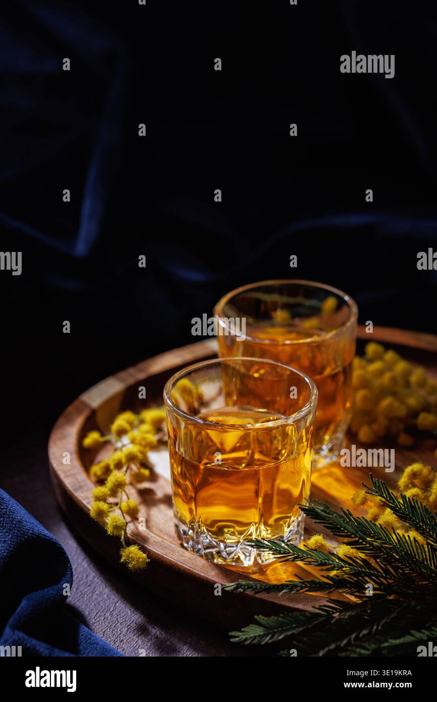 Zwei Gläser Whiskey oder Brandy mit Mimosa-Blumen auf Holztablett, Frühlingsgetränk-Konzept Stockfoto