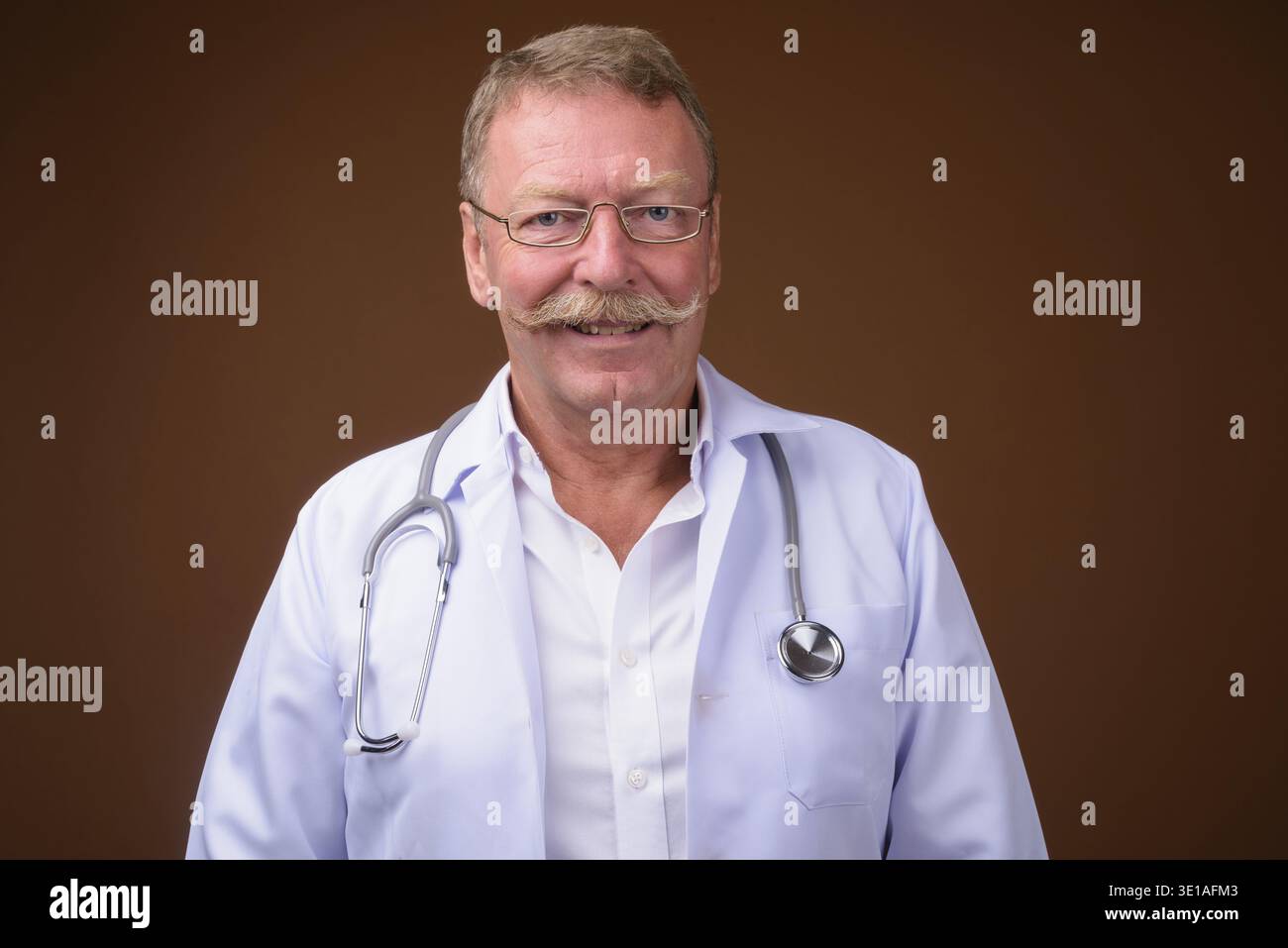 Studio-Aufnahme eines gutaussehenden Seniorendoktors mit Schnurrbart, der eine Brille vor braunem Hintergrund trägt Stockfoto