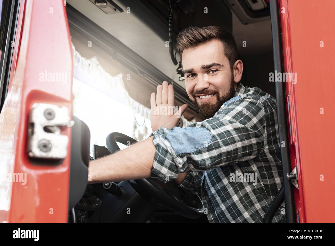 Junger, lächelnder Lkw-Fahrer, der im Truck sitzt und mit der Hand winkt Stockfoto