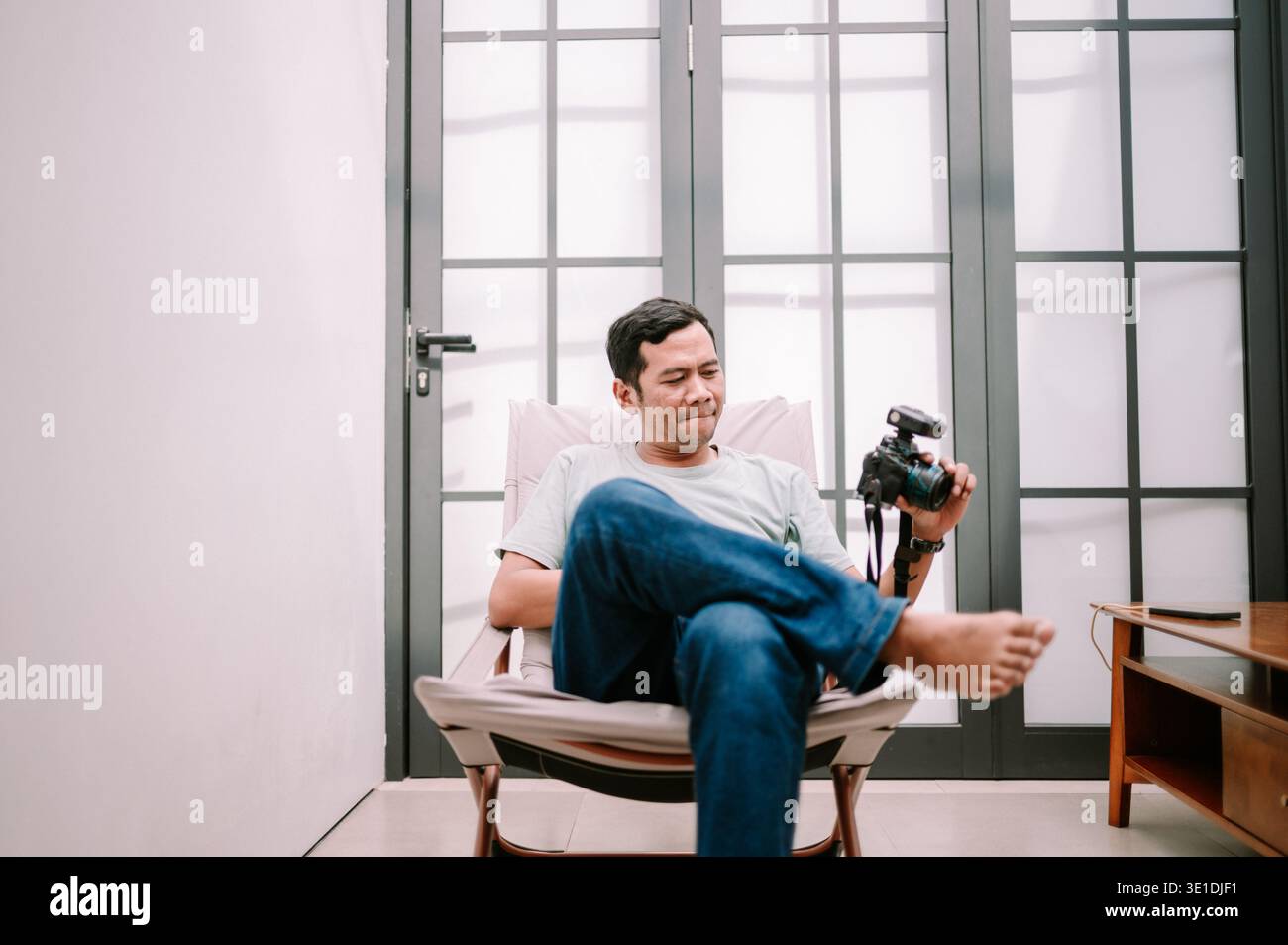 Ein entspannter Mann in lässiger Kleidung sitzt bequem und betrachtet Bilder auf seiner Digitalkamera mit einem externen Blitz und genießt sein Hobby zu Hause Stockfoto