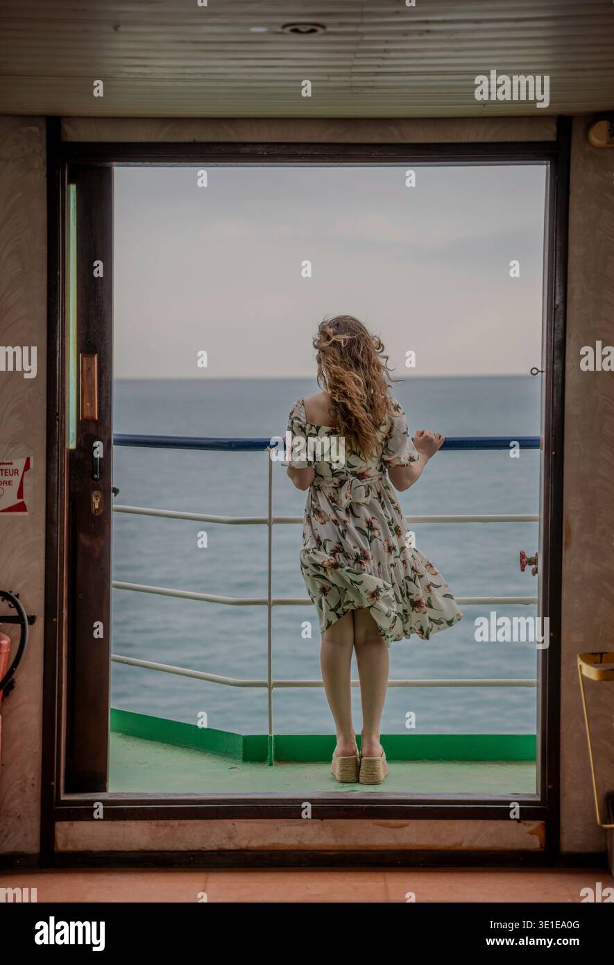 Eine Frau in einem Blumenkleid steht am offenen Geländer eines Fährdecks und blickt auf das ruhige Mittelmeer während einer Überfahrt. Stockfoto