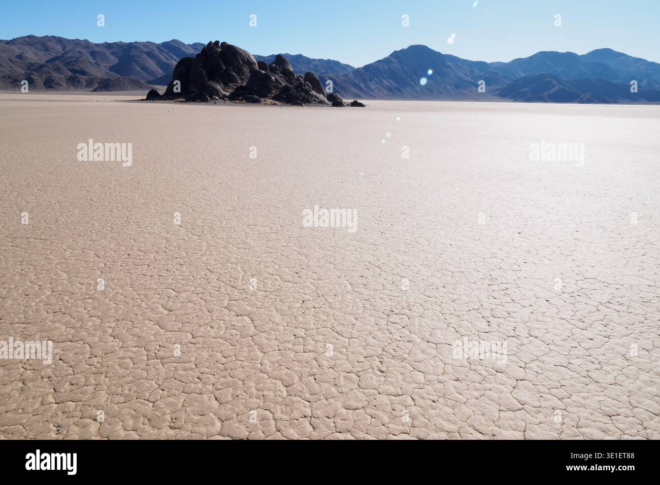 Eine riesige, zerrissene Schlammfläche erstreckt sich zu einer Ansammlung dunkler, felsiger Formationen unter einem klaren blauen Himmel. Die Landschaft wirkt trocken und einsam, mit d Stockfoto