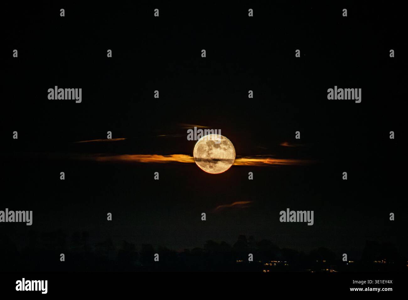 Ein Vollmond ist hinter Wolken zu sehen, die einen orangen Schein über den Horizont werfen. Der Mond wird teilweise von den Wolken verdeckt, was ein dramatisches Bild erzeugt Stockfoto