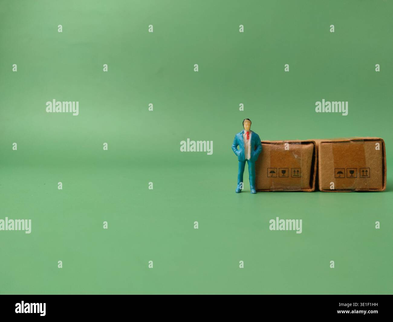 Kleine Geschäftsmannsfigur, die neben zwei kleinen Papppaketen auf einem grünen Hintergrund steht. Stockfoto