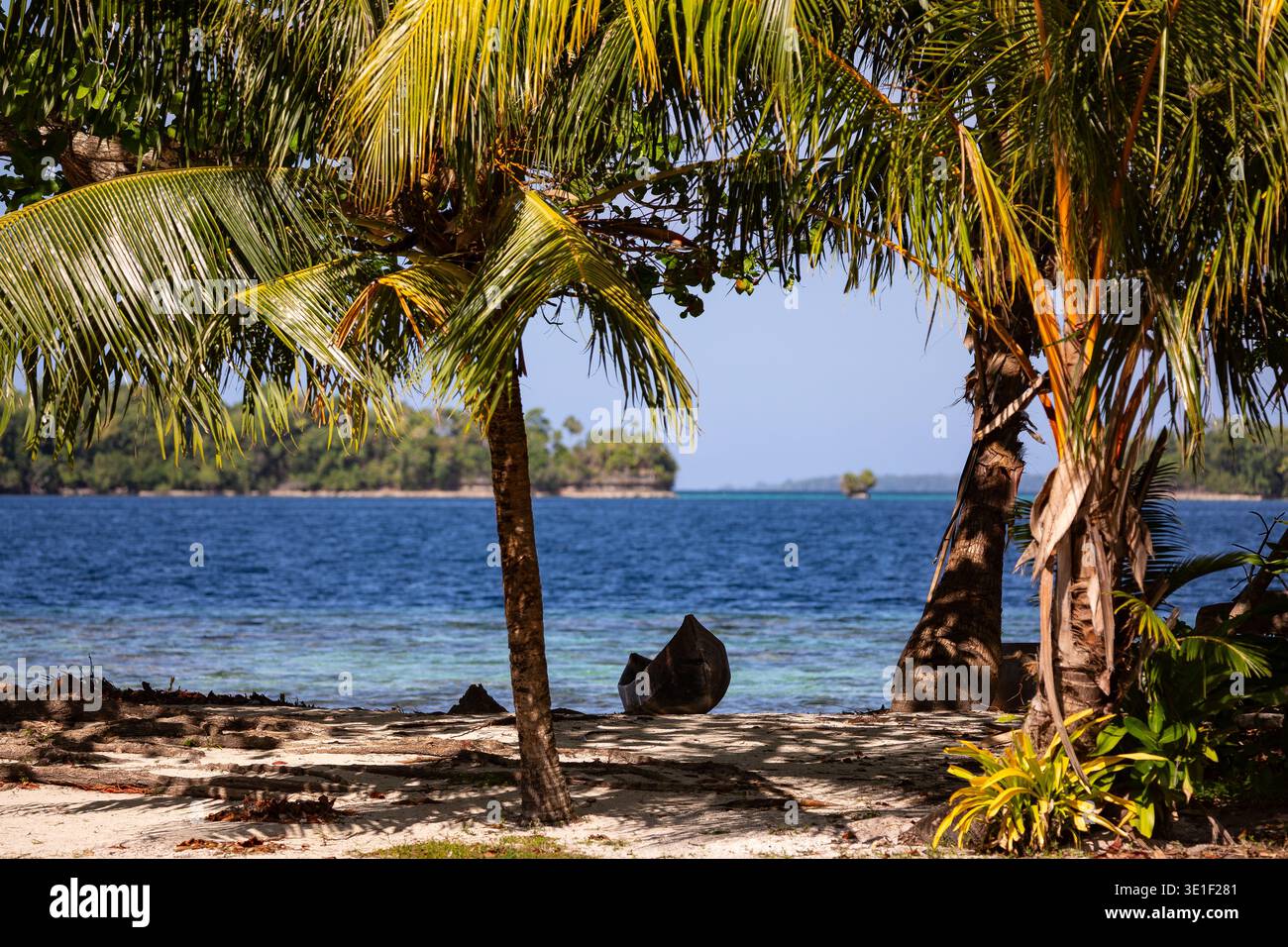 Ein Kanu liegt an einem Sandstrand im Schatten von Palmen, mit dem blauen Meer und den üppigen grünen Inseln im Hintergrund. Stockfoto
