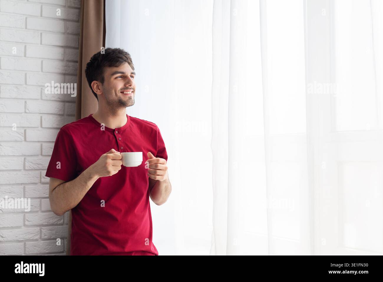 Glücklicher Mann, der eine Tasse Kaffee am Fenster hält Stockfoto