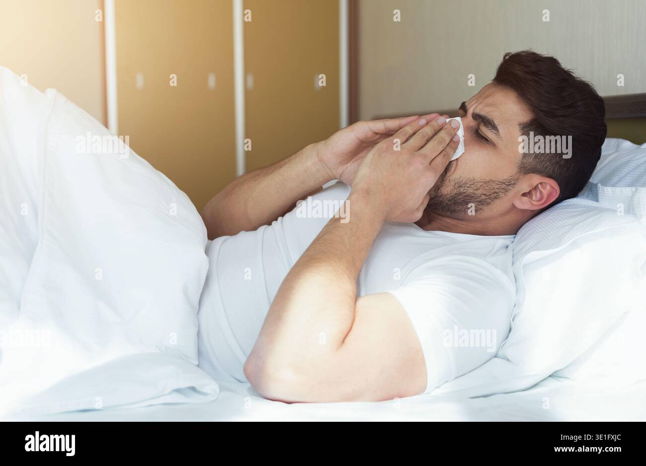Junger Mann niest und bedeckt die Nase mit auf dem Bett liegendem Gewebe Stockfoto
