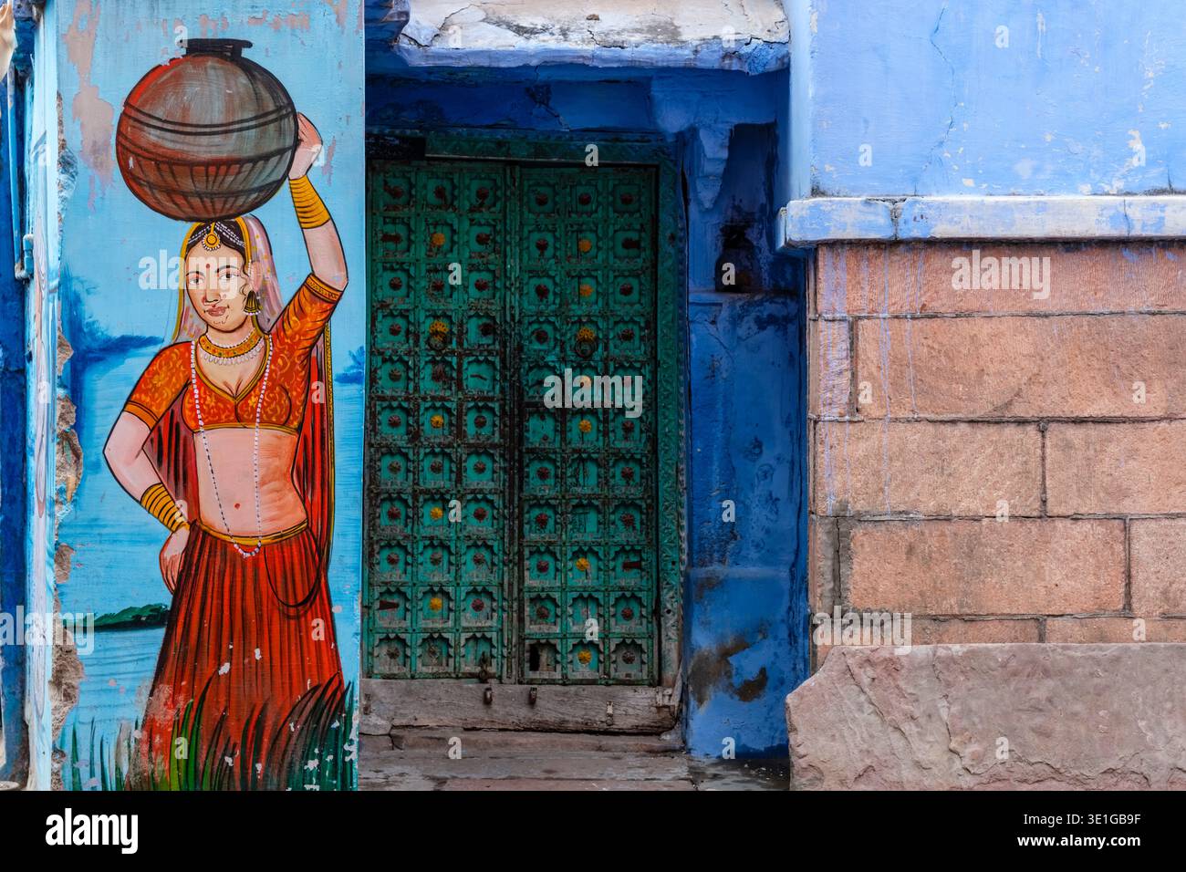 Ein traditionelles Wandbild einer Frau mit einem Topf ziert eine blaue Wand neben einer aufwendig geschnitzten grünen Holztür in Jodhpur. Stockfoto