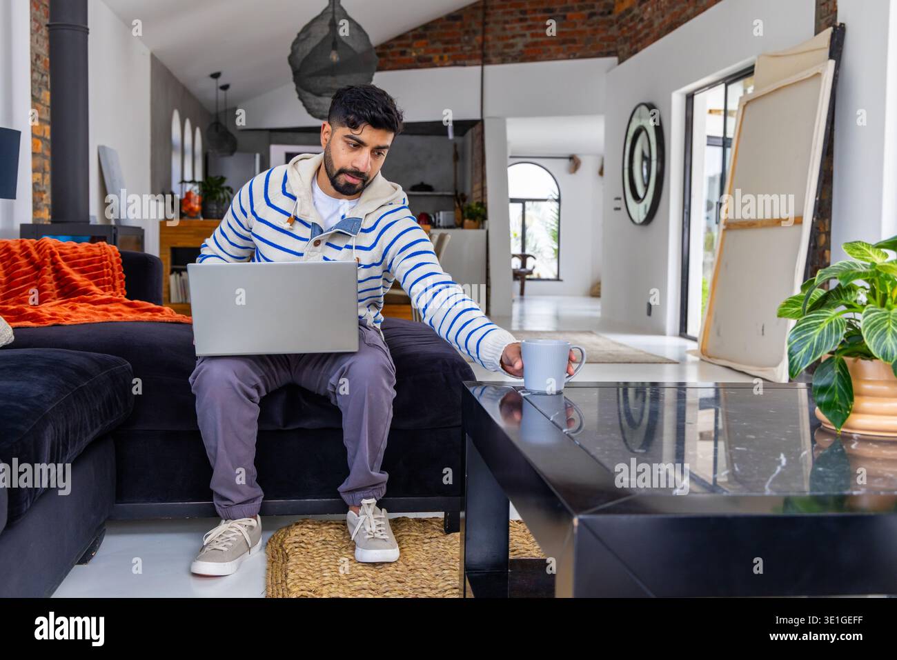 Mann, der auf einem separaten Sofa im Wohnzimmer sitzt und nach Becher greift, während er am Laptop arbeitet Stockfoto