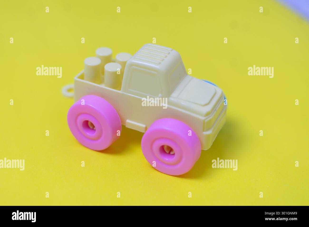 Ein kleiner, cremefarbener Spielzeugwagen aus Kunststoff mit hellrosafarbenen Rädern ist auf einem leuchtend gelben Hintergrund positioniert und eignet sich perfekt für Kinderspiele und Fantasie Stockfoto