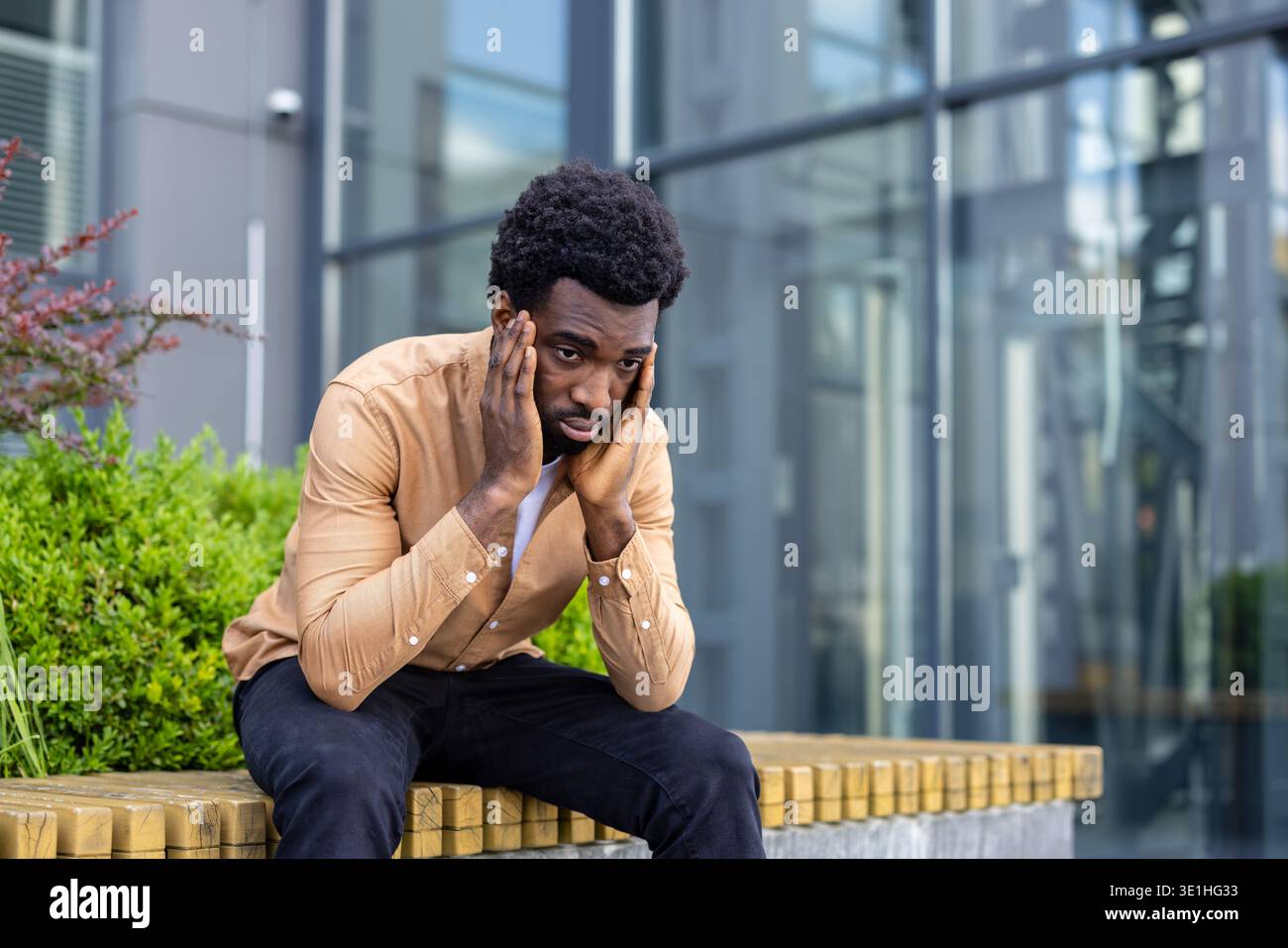 Junger Mann, der sich überwältigt und unglücklich fühlt, allein draußen sitzt und die Hände an den Kopf gedrückt hat, was auf Stress, Enttäuschung oder mentalen Kampf hindeutet Stockfoto
