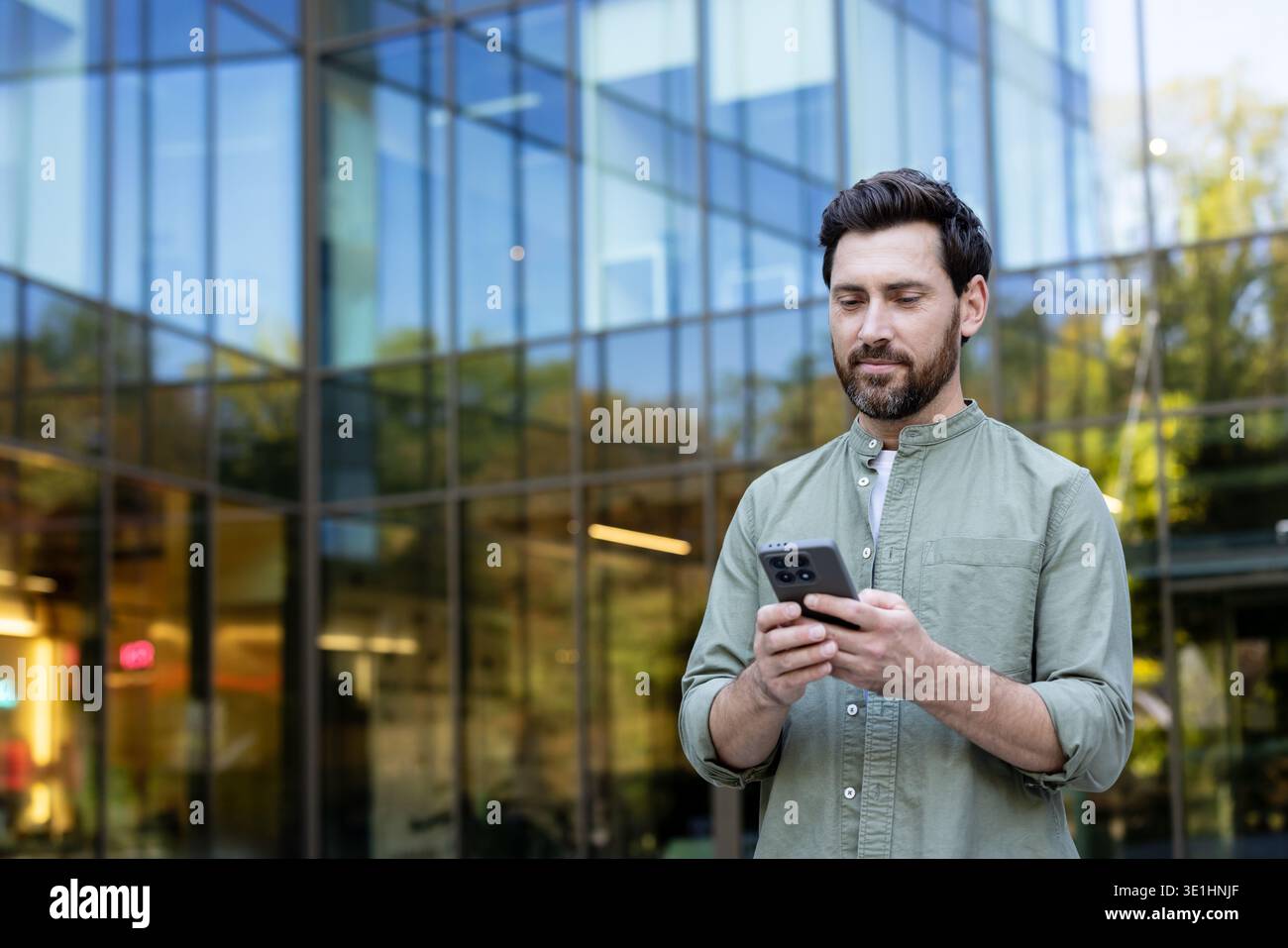 Junger bärtiger Mann, der draußen vor einem Glasgebäude stand, konzentrierte sich auf sein Smartphone für Messaging, Surfen und mobile Konnektivität in einem städtischen Geschäftsumfeld Stockfoto