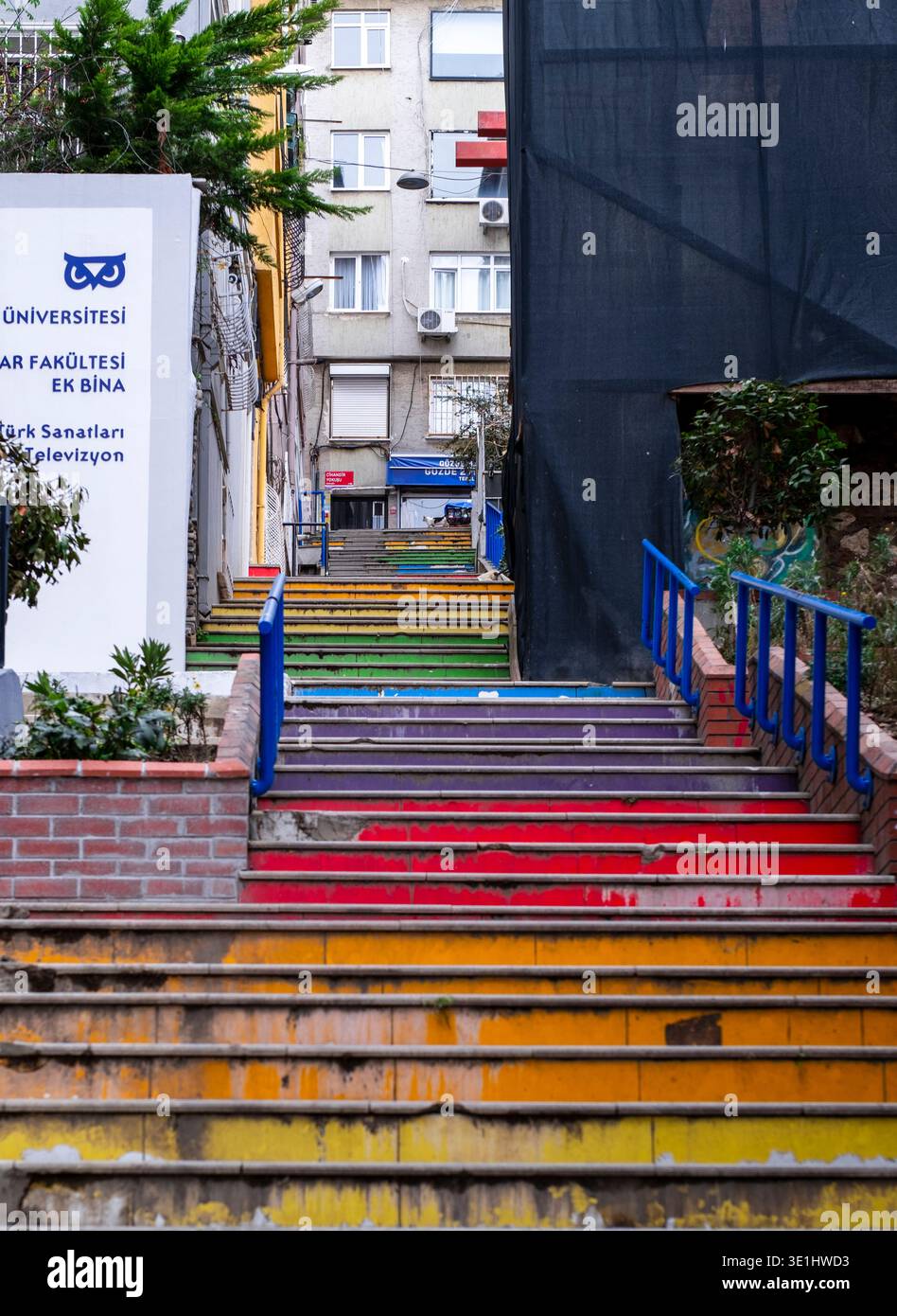 Regenbogenfarbene Betontreppen mit blauen Geländern steigen zu Wohngebäuden und einem Universitätsschild auf. Cihangir, Istanbul, Türkei Stockfoto