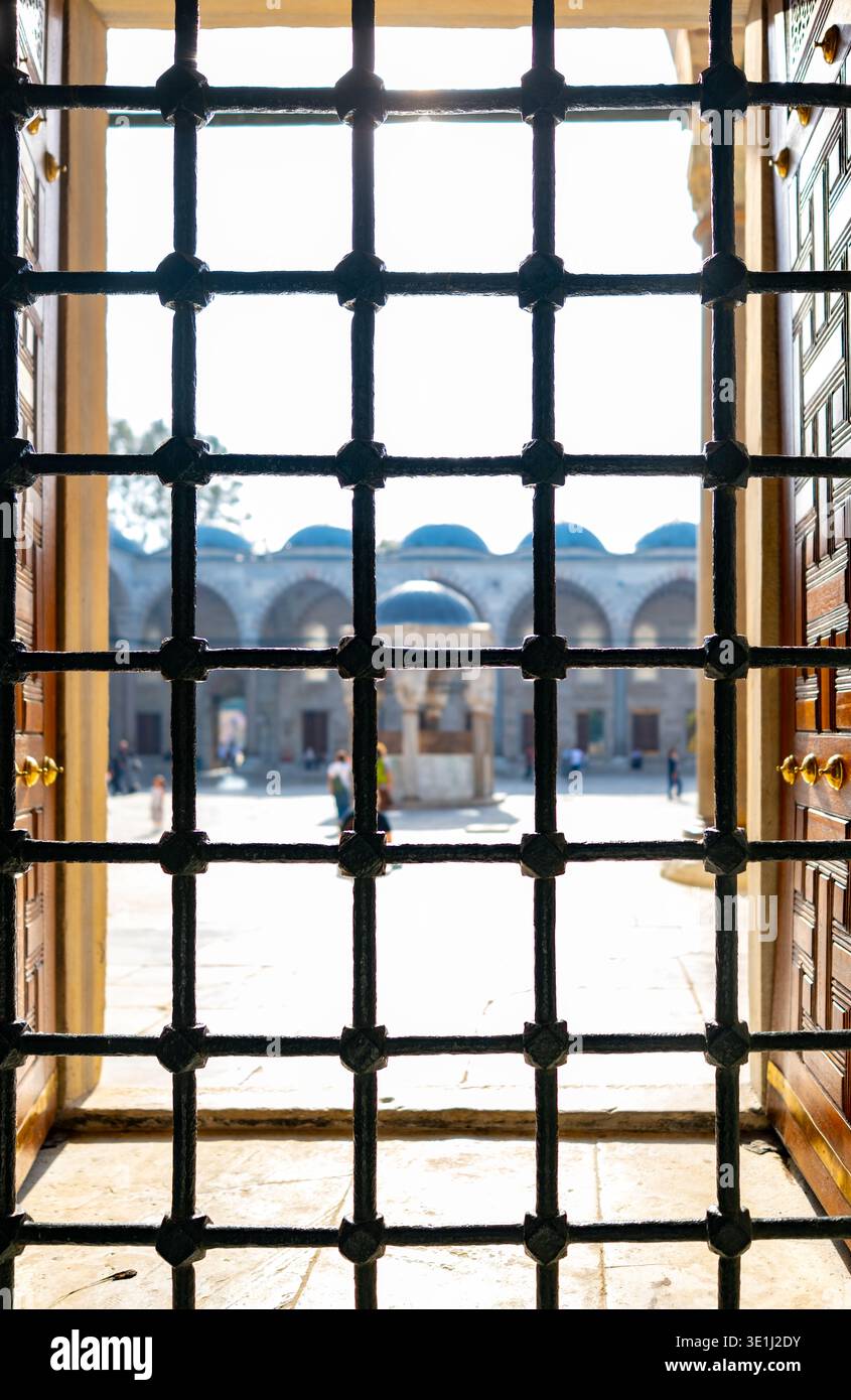 Ein Blick durch dekorative schmiedeeiserne Fensterstangen zeigt einen sonnendurchfluteten historischen europäischen Innenhof mit klassischer Bogenarchitektur und Spaziergänge Stockfoto