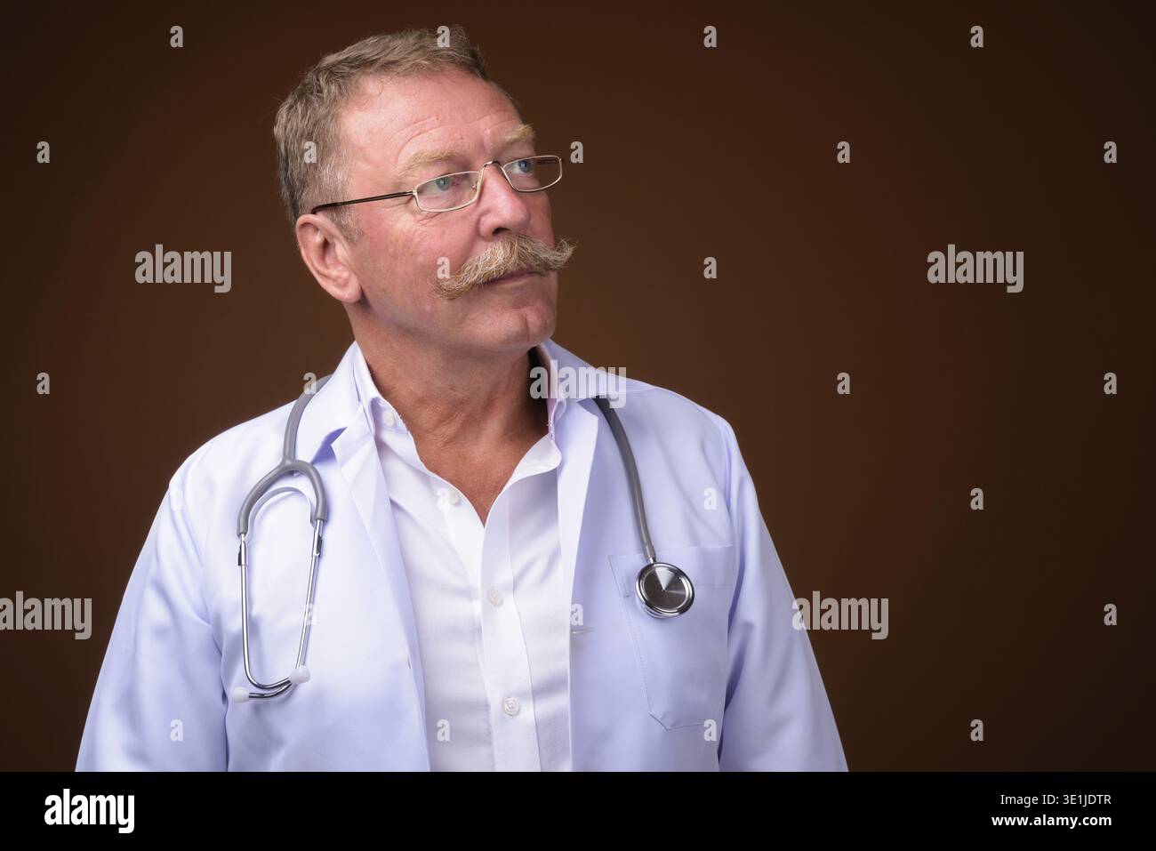 Studio-Aufnahme eines gutaussehenden Seniorendoktors mit Schnurrbart, der eine Brille vor braunem Hintergrund trägt Stockfoto