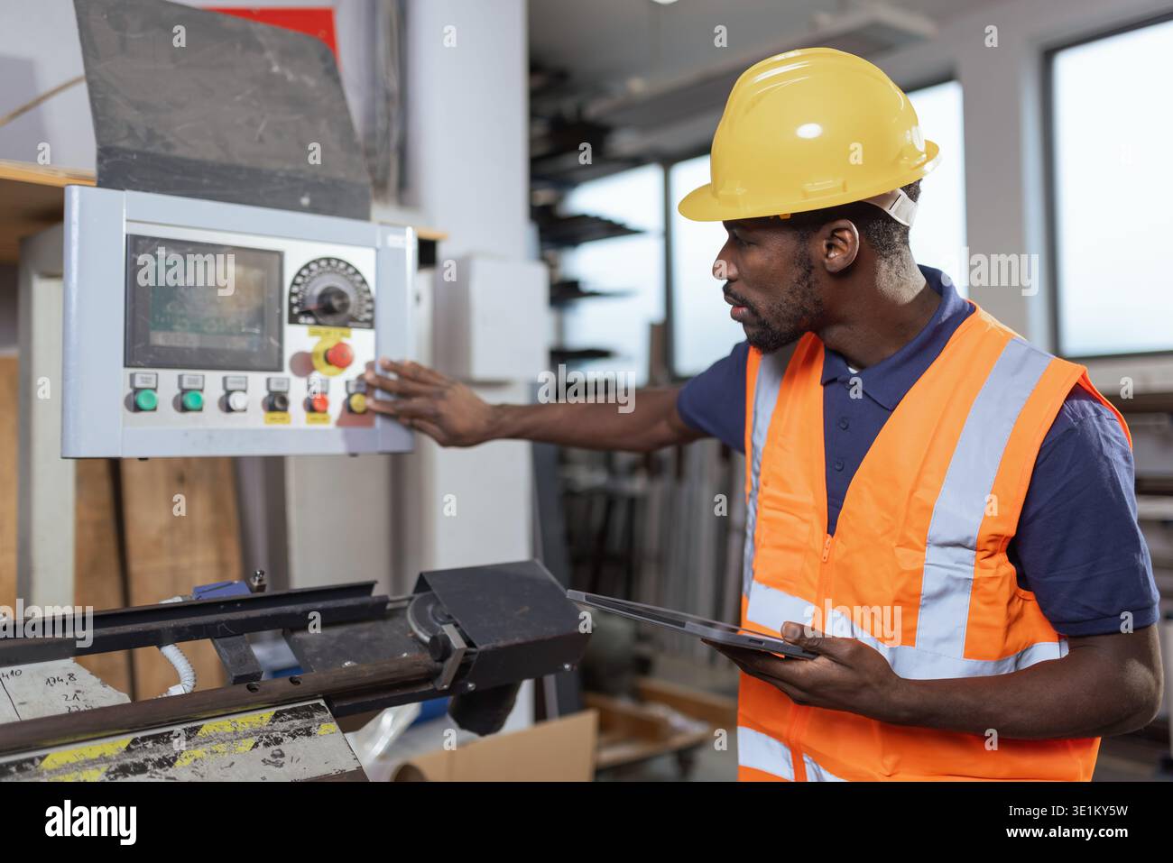 Mitarbeiter der Metallindustrie mit gelbem Schutzhelm überwacht den automatisierten Systembetrieb und nimmt Anpassungen per Tablet vor, um Prozesse zu optimieren. Stockfoto