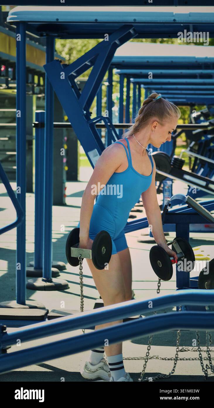 Eine Frau in einem blauen Sportanzug, die sich auf ihr Workout konzentriert, hebt Kurzhanteln in einem Fitnessstudio im Freien. Dieses Foto zeigt, wie wichtig es ist, Stärke zu integrieren Stockfoto