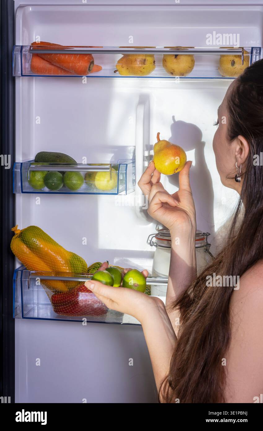 Junge Frau, die den Kühlschrank öffnet und hineinblickt, während sie frisches Obst auswählt. Konzept von gesunder Ernährung, Ernährung und Küchenalltag Stockfoto