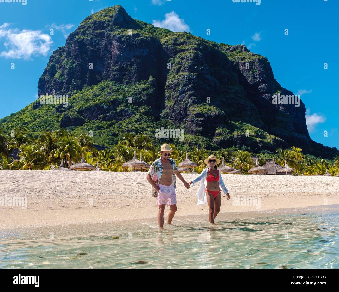 In der atemberaubenden Landschaft von Mauritius spazieren ein Paar Hand in Hand am kristallklaren Wasser entlang. Der lebhafte grüne Berg ragt majestätisch hinter sich, perfekt für einen romantischen Kurzurlaub. Stockfoto