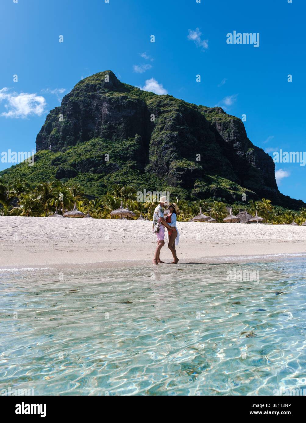 Goldener Sand und klares Wasser schaffen eine ruhige Atmosphäre, während ein Paar einen gemütlichen Spaziergang am Strand von Mauritius genießt, umgeben von beeindruckenden Berglandschaften von Le Morne Mauritius Stockfoto
